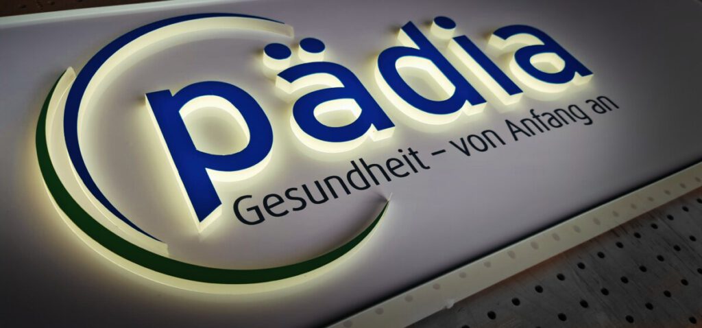 Beleuchtete Acrylglas-Leuchtbuchstaben 'Pädia' mit dem Slogan 'Gesundheit - von Anfang an' auf weißem Hintergrund.